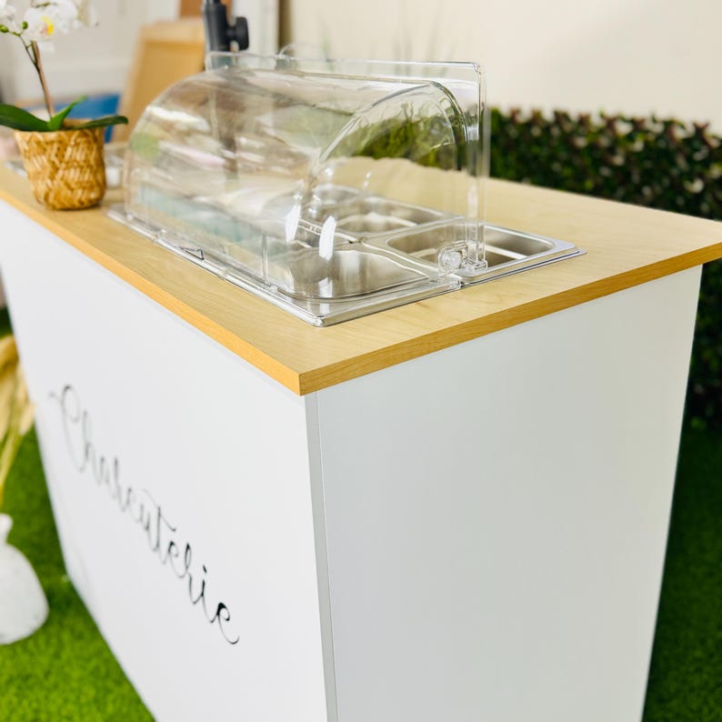 May include: A white charcuterie display table with a light wood top and a clear plastic food cover. The table has a stainless steel food tray and the word "Charcuterie" is written on the side in cursive.