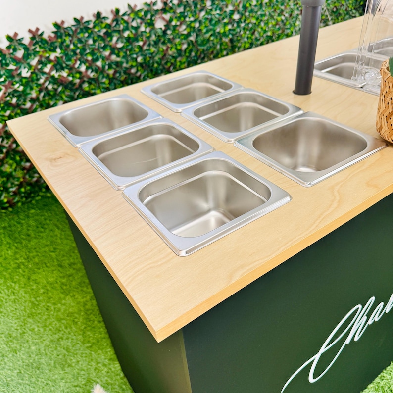 Pu&ograve; includere: Un'isola di servizio in legno con sei contenitori per alimenti in acciaio inossidabile. L'isola ha un piano in legno chiaro e una base verde scuro. Sono visibili anche un contenitore trasparente e un cestino di vimini.