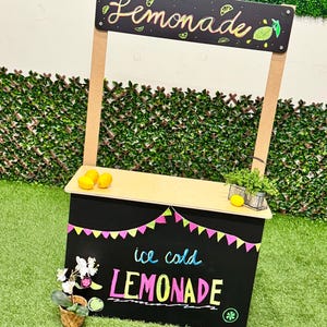 May include: A small, wooden lemonade stand with a black chalkboard front and top. The sign above reads "Lemonade" in cursive. The chalkboard advertises "ice cold LEMONADE" in colorful chalk. The stand is decorated with lemons and a small plant.