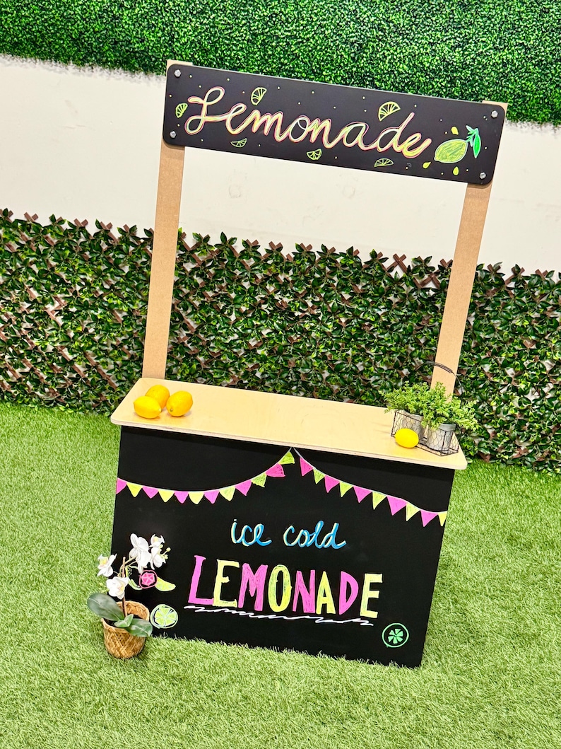 May include: A small, wooden lemonade stand with a black chalkboard front and top. The sign above reads "Lemonade" in cursive. The chalkboard advertises "ice cold LEMONADE" in colorful chalk. The stand is decorated with lemons and a small plant.