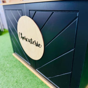 May include: A black wooden cabinet with a geometric design on the front. A round wooden sign with the word "Charcuterie" is attached to the cabinet.