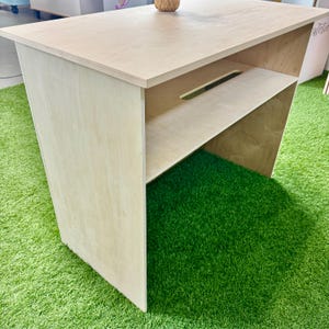 Pu&ograve; includere: Un tavolo in legno color chiaro con piano rettangolare e ripiano. Il tavolo ha un design semplice con linee pulite. Il tavolo &egrave; appoggiato su una superficie verde.