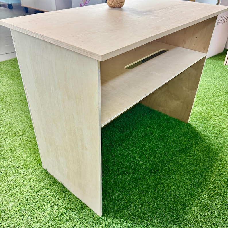 Pu&ograve; includere: Un tavolo in legno color chiaro con piano rettangolare e ripiano. Il tavolo ha un design semplice con linee pulite. Il tavolo &egrave; appoggiato su una superficie verde.