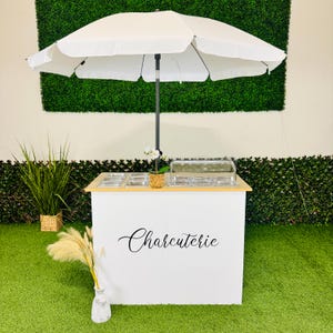 May include: A white charcuterie cart with a wooden top and a large white umbrella. The cart has the word "Charcuterie" in elegant script. The cart is on a green surface with plants in the background.