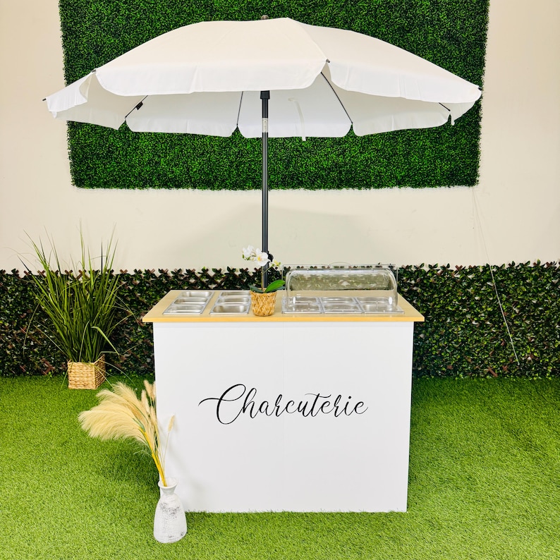 May include: A white charcuterie cart with a wooden top and a large white parasol. The cart has the word "Charcuterie" in elegant script. The cart is on a green surface with plants in the background.