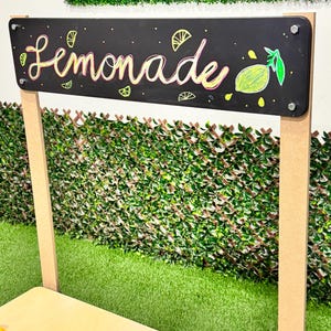 May include: A black chalkboard sign with the word "Lemonade" in colorful script, adorned with lemon illustrations. The sign is mounted on a wooden frame, set against a backdrop of green foliage and artificial grass.