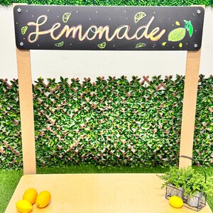 May include: A wooden lemonade stand with a black chalkboard sign that reads "Lemonade" in colorful script. The stand has a light wood countertop, lemons, and decorative plants. A green hedge backdrop completes the scene.