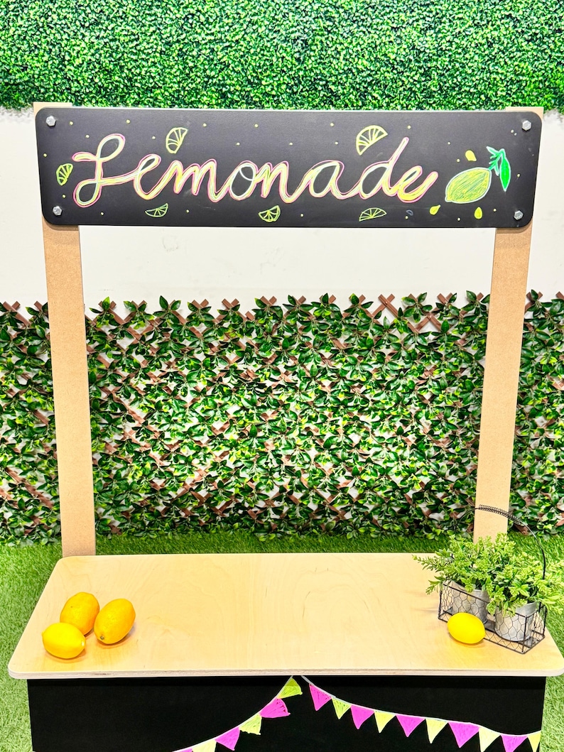 May include: A wooden lemonade stand with a black chalkboard sign that reads "Lemonade" in colorful script. The stand has a light wood countertop, lemons, and decorative plants. A green hedge backdrop completes the scene.