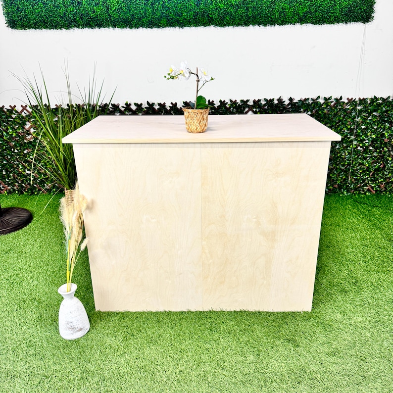 May include: A light-coloured wooden bar with a flat top and angled sides. A small potted orchid sits on top. The bar is set on a green surface with decorative plants and a white wall in the background.