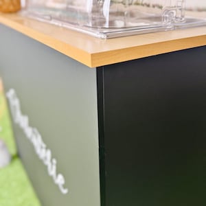 Pu&ograve; includere: Primo piano di una vetrina con un piano in legno chiaro e un pannello laterale nero. Il pannello laterale &egrave; diviso in due sezioni, una grigio chiaro e l'altra nera. Il testo "The Barbie" &egrave; visibile sulla sezione grigia.