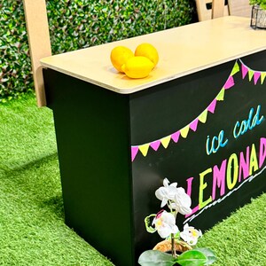 May include: A black lemonade stand with a light wood top. The stand has colorful text that says "ice cold LEMONADE". Three yellow lemons sit on top. A small potted orchid is in front of the stand.