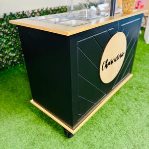 May include: A black wooden food cart with a wooden top and a round wooden sign that says "Charcuterie". The cart has a geometric pattern on the front and is on wheels.