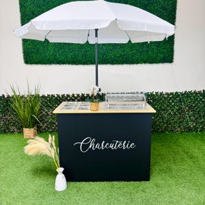 May include: A black charcuterie cart with a white umbrella. The cart has a wooden countertop with food containers and a clear display case. The word "Charcuterie" is written in white script on the front.