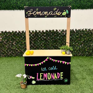 May include: A wooden lemonade stand with a black base and a light-colored countertop. The sign reads "Lemonade" in cursive and "ice cold LEMONADE" in colorful lettering. The stand is decorated with lemons and a garland of pink and green flags.