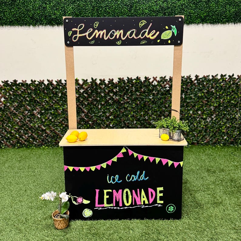 May include: A wooden lemonade stand with a black base and a light-colored countertop. The sign reads "Lemonade" in cursive and "ice cold LEMONADE" in colorful lettering. The stand is decorated with lemons and a garland of pink and green flags.