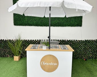 May include: A white charcuterie cart with a white umbrella. The cart has a wooden countertop with stainless steel containers and a clear cover. The cart has a wooden circle with the word "Charcuterie" on it.