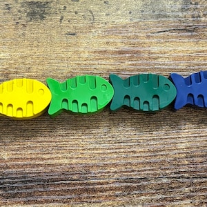 Könnte beinhalten: Eine Reihe von neun bunten fischförmigen Buntstiften, die in einem Regenbogenmuster auf einer Holzoberfläche angeordnet sind. Die Stifte sind rot, orange, gelb, grün, blau, lila, rosa und schwarz.