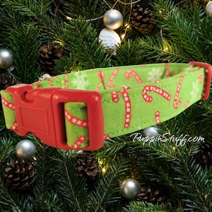 Può includere: Un collare per animali in tessuto verde con un motivo ripetuto di bastoncini di zucchero rossi e bianchi e fiocchi di neve bianchi. Il collare include una fibbia in plastica rossa a sgancio laterale e un cursore, con il testo RuppinStuff.com.