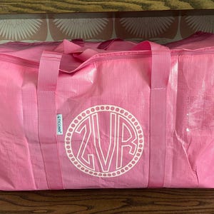 Op de afbeelding: Een grote, roze draagtas met ritssluiting en stevige handvatten. De tas heeft een wit monogramontwerp in een cirkelvormig frame. De tas is gemaakt van een duurzaam, getextureerd materiaal.