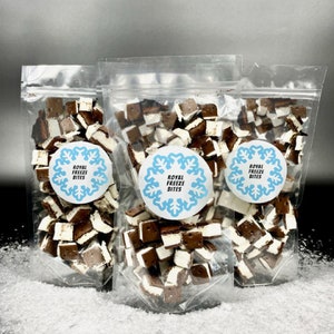 May include: Three clear plastic bags filled with bite-sized chocolate and vanilla treats. Each bag has a circular label with the text "ROYAL FREEZE BITES" in blue. The treats are arranged on a surface that resembles snow.