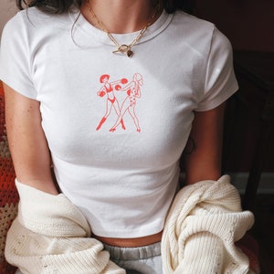 May include: A white cropped t-shirt featuring a red graphic of two women boxing. The shirt has a gold chain necklace with a heart pendant.