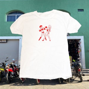 May include: White t-shirt featuring a red graphic of two women boxing. The women are wearing boxing gloves and shorts.