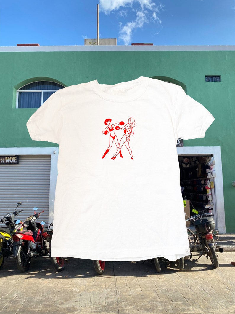 May include: White t-shirt featuring a red graphic of two women boxing. The women are wearing boxing gloves and shorts.