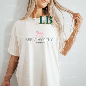 May include: White t-shirt with a pink ribbon and the text "SHE IS WORTHY" with the scripture reference "PROVERBS 31:10" underneath.