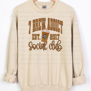 May include: A beige long-sleeve crewneck sweatshirt featuring a brown graphic print that reads 7 BREW ADDICT EST. 2017 Social club, with an illustration of a coffee cup with whipped cream in the center of the text.