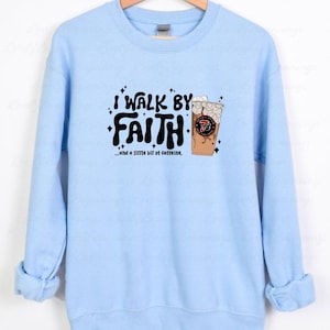 May include: Light blue sweatshirt with the text "I Walk By Faith...and a little bit of caffeine." The graphic includes a coffee drink with ice and the number 73. The sweatshirt is hanging on a wooden hanger.