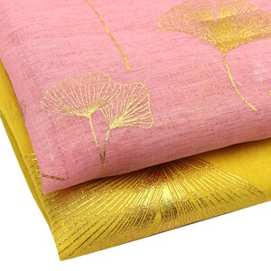 May include: Two folded pieces of fabric stacked together. The top layer is a light pink textile featuring a metallic gold ginkgo leaf pattern. The bottom layer is a vibrant yellow textile with a large, detailed metallic gold ginkgo leaf print.