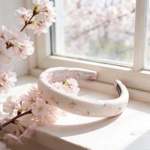 以下が含まれることがあります： クリーム色のパッド入りヘアバンド。ピンクと白の刺繍の花柄が施されています。ヘアバンドは、ピンクの桜の枝の隣の窓枠に置かれ、春らしい雰囲気を醸し出しています。