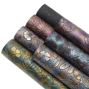 May include: Five rolls of iridescent faux leather with an embossed floral and butterfly design. The rolls are arranged in a stack, with the darkest roll at the top and the lightest roll at the bottom. The colors range from deep purple to gold.