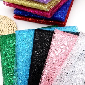May include: A collection of colorful glitter fabric squares in various shades, including green, blue, black, pink, and silver. Additional squares in red, blue, pink, gold, and purple are stacked in the background. The fabric has a mesh-like texture.