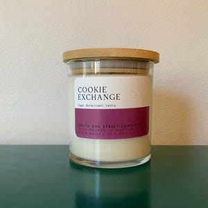 Può includere: Un barattolo di vetro per candele con un coperchio di legno. La candela è bianca e ha un'etichetta che recita "COOKIE EXCHANGE" con il profumo "Sugar Buttercream, Vanilla". L'etichetta riporta anche "South Ann Street Candle Co." e "Hand Poured in Maryland".