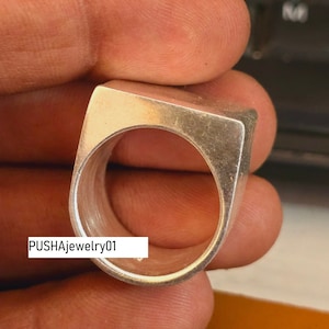 May include: A silver ring with a square face and a smooth, polished finish. The ring is held in a hand, and the text "PUSHAjewelry01" is visible on the band.