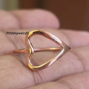 May include: A rose gold heart-shaped ring with a delicate open design. The ring is shown on a finger.