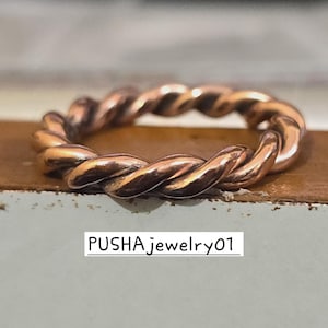 Könnte beinhalten: Ein Kupferring mit einem gedrehten Design. Der Ring besteht aus einem einzigen Stück Kupferdraht, das zu einem seilartigen Effekt verdreht wurde. Der Ring ist ein schlichtes und elegantes Design, das ein tolles Geschenk für einen besonderen Menschen wäre.