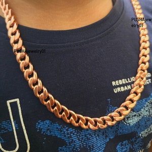 May include: A copper chain necklace with a curb link design. The chain is thick and has a shiny finish. The necklace is worn by a person with a dark blue shirt.