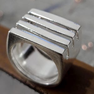 Peut inclure: Une bague en argent au design rectangulaire. La bague présente quatre barres parallèles sur le dessus, créant un look géométrique moderne. L'anneau est lisse et poli, reflétant la lumière.