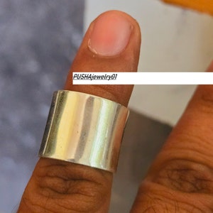 May include: A silver band ring with a wide, flat top. The ring is worn on a finger.
