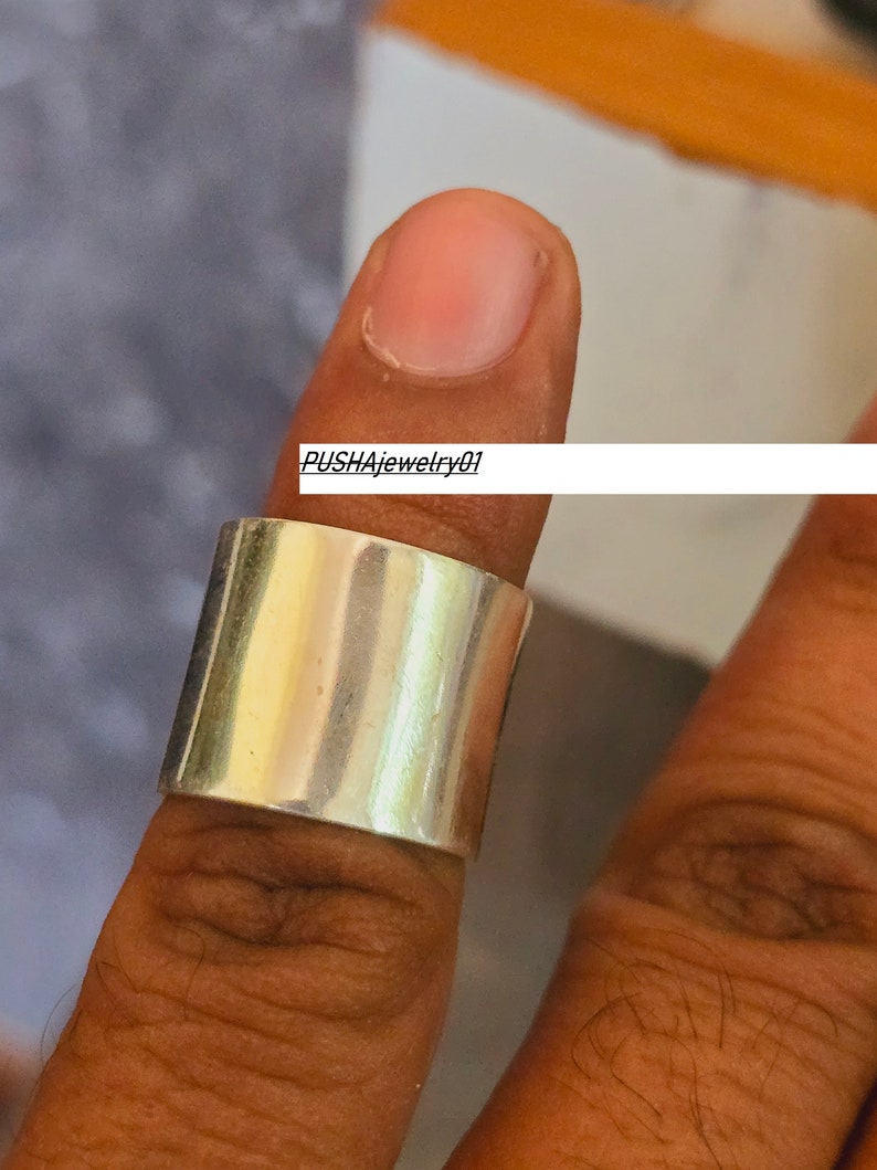 May include: A silver band ring with a wide, flat top. The ring is worn on a finger.