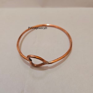 May include: A simple copper bangle bracelet with a heart-shaped cutout. The bracelet is on a white background.  PUSHAJewelry01