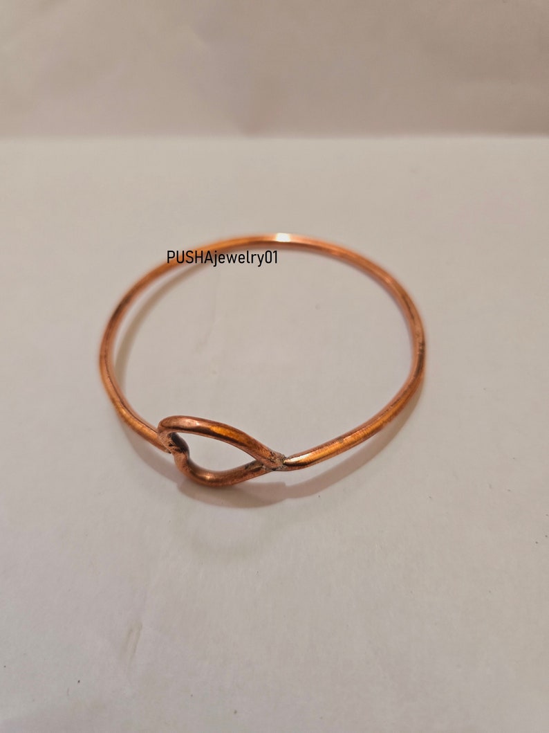 May include: A simple copper bangle bracelet with a heart-shaped cutout. The bracelet is on a white background.  PUSHAJewelry01