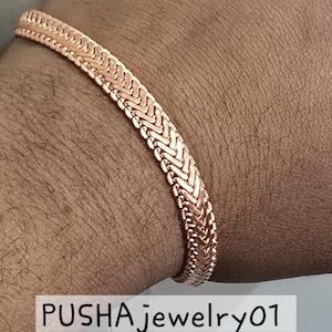 Peut inclure: Un bracelet en chaîne or rose avec un motif tissé. Le bracelet est porté au poignet.