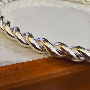 May include: A silver twisted bangle bracelet with a polished finish. The bracelet is thick and has a rope-like design.