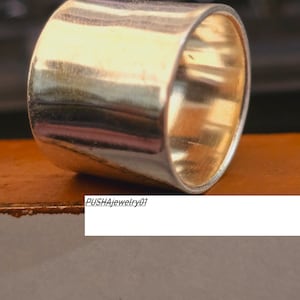 May include: A silver ring with a wide, flat band. The ring is smooth and shiny, and it has a slightly rounded edge. The ring is sitting on a brown surface.  PUSHAjewelry01