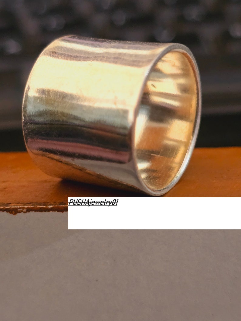 May include: A silver ring with a wide, flat band. The ring is smooth and shiny, and it has a slightly rounded edge. The ring is sitting on a brown surface.  PUSHAjewelry01