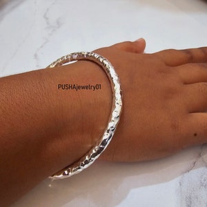 May include: A silver hammered bangle bracelet. The bracelet is thick and has a textured surface. The bracelet is worn on a person's wrist.
