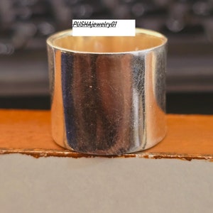 May include: A silver ring with a wide band. The ring is smooth and shiny. The text "PUSHAjewelry01" is visible on the ring.